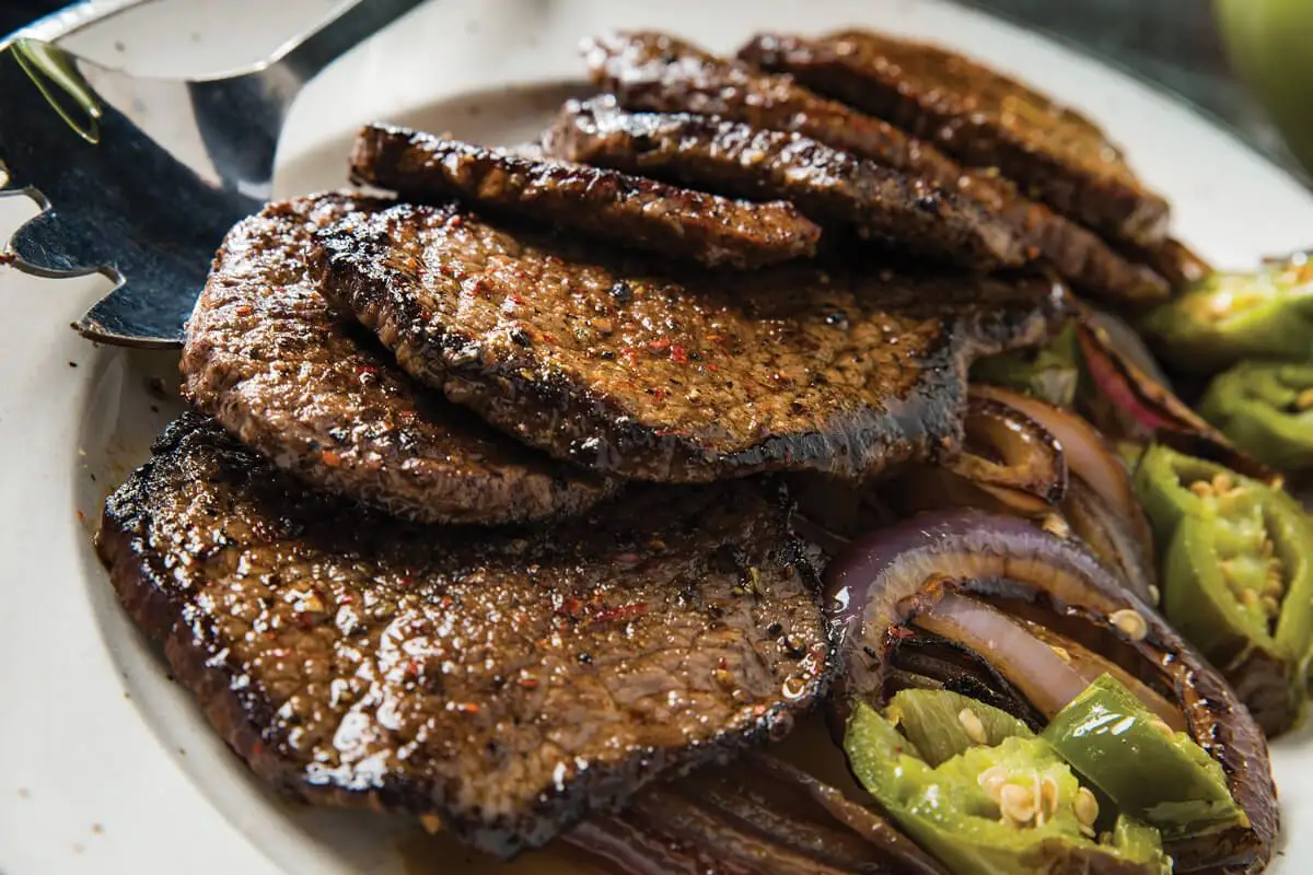 Two browned eye of round steaks resting on a plate near a stove, with a skillet in the background