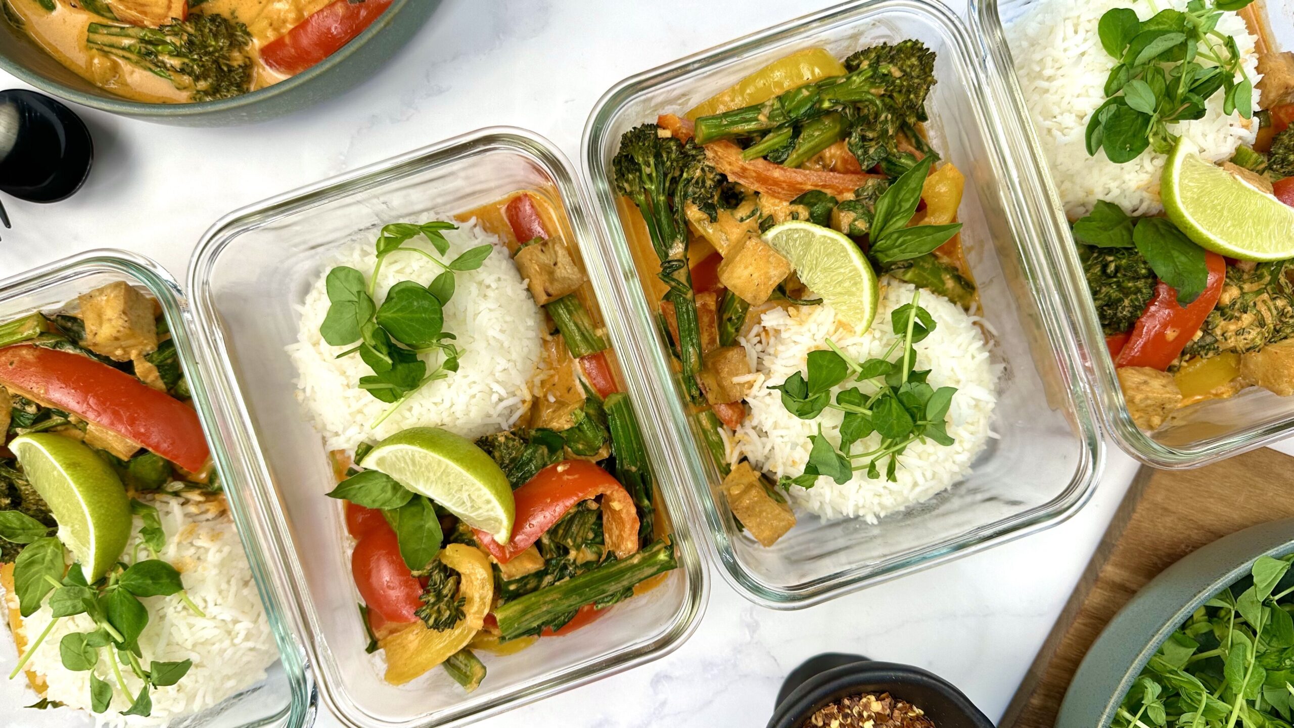 Two glass meal prep containers filled with Thai red curry and jasmine rice on a kitchen counter, lids set nearby
