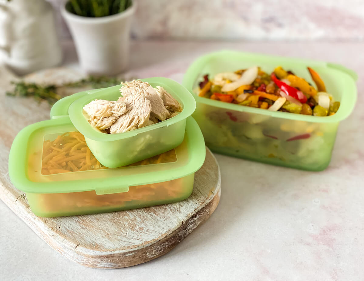 Two glass meal prep containers filled with cowboy stew, lids off, sitting on a kitchen counter with a refrigerator door open in the background