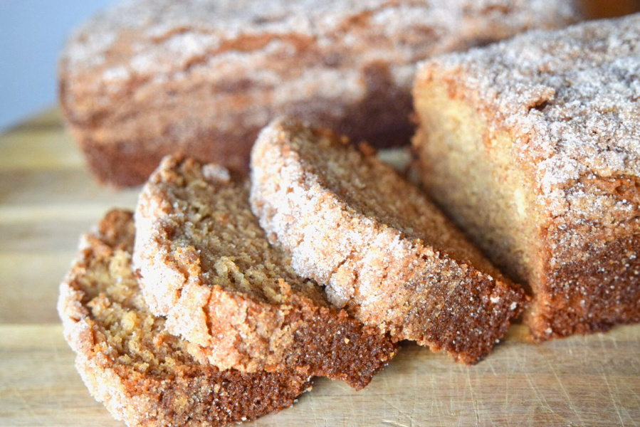 Two golden loaves of Amish Friendship Bread on a wooden cutting board with cinnamon sugar sprinkled on top, one loaf sliced to show a tender cinnamon swirl