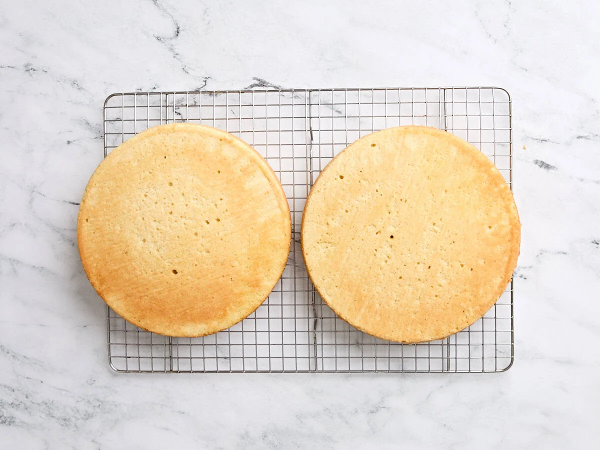 Two golden vanilla cake layers cooling on wire racks on a kitchen counter, parchment paper set aside