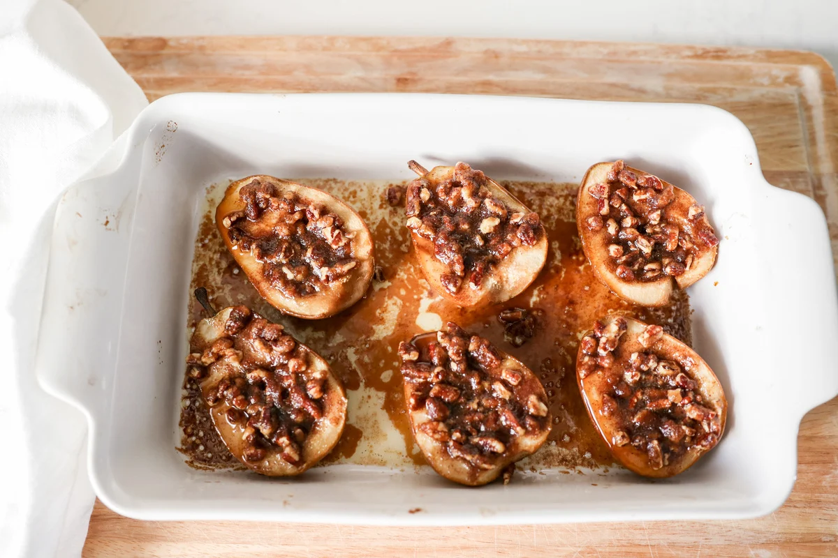 Two halved baked pears on a small white plate, glossy with brown sugar syrup and a scoop of vanilla ice cream beside them on a wooden kitchen table