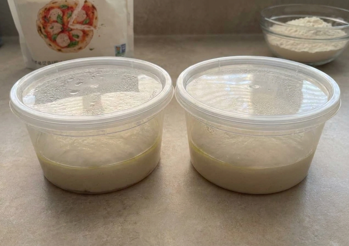 Two lightly oiled pizza dough balls resting in covered deli containers on a kitchen counter, ready for cold fermentation