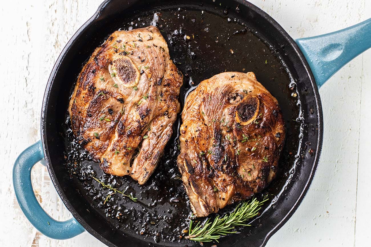 Two mutton shoulder chops searing in a cast iron skillet with browned edges forming and oil shimmering on a home stovetop