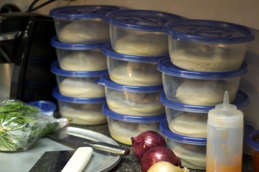 Two oiled pizza dough balls in clear lidded containers resting on a kitchen counter, ready for the refrigerator