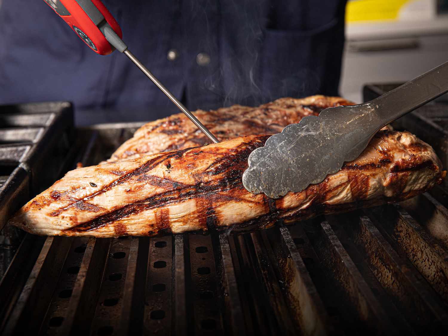 Two pork tenderloins searing on a hot outdoor grill with visible grill marks and a pair of tongs turning one tenderloin