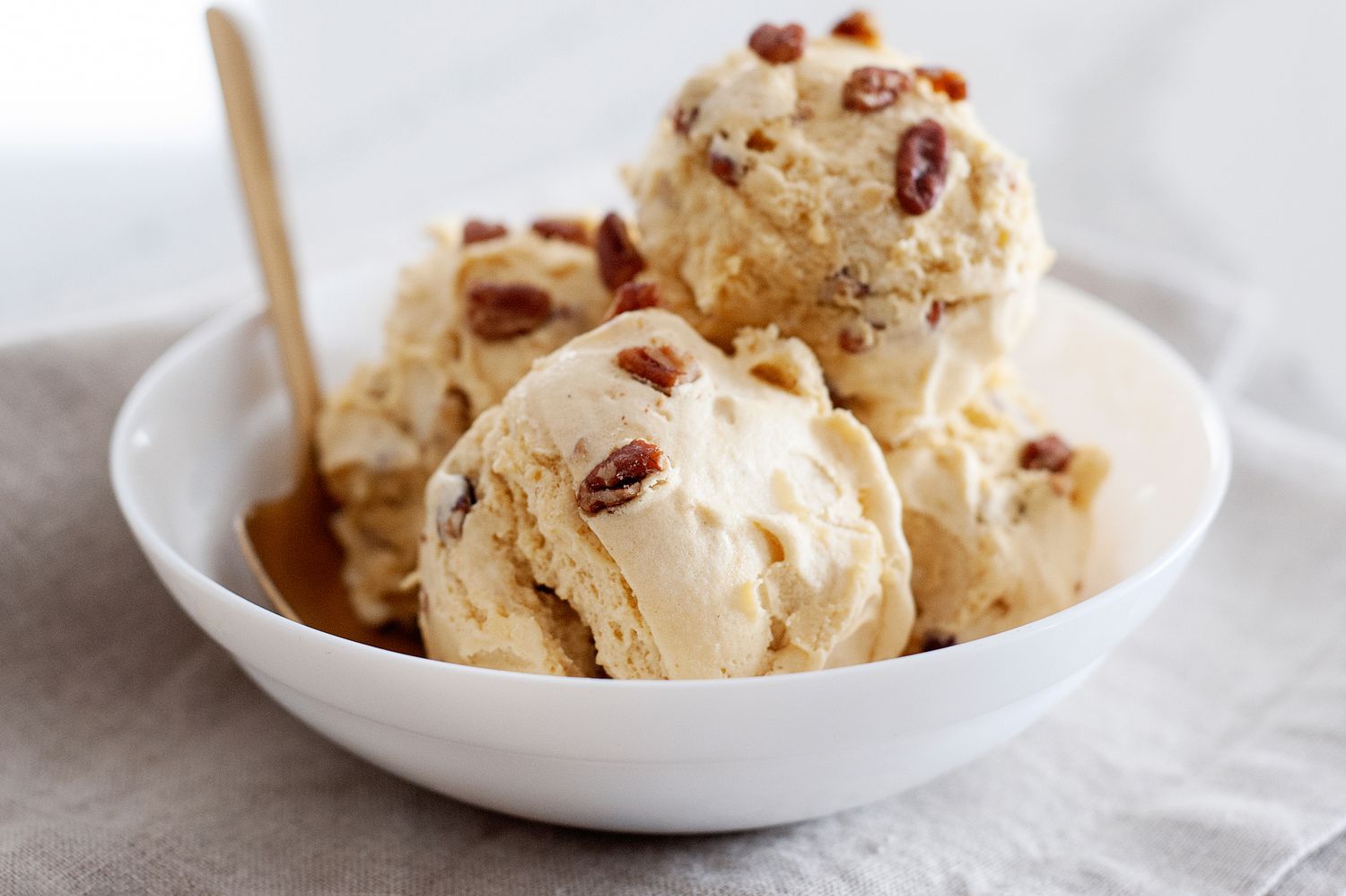 Two scoops of butter pecan ice cream in a ceramic bowl with toasted pecans scattered on top, sitting on a light kitchen table with a spoon beside it