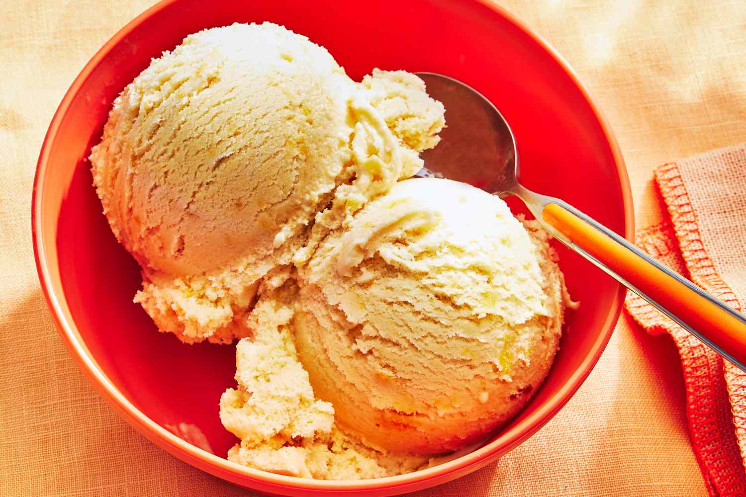 Two scoops of homemade peach ice cream in a ceramic bowl with visible peach pieces, set on a light kitchen table with a spoon beside it