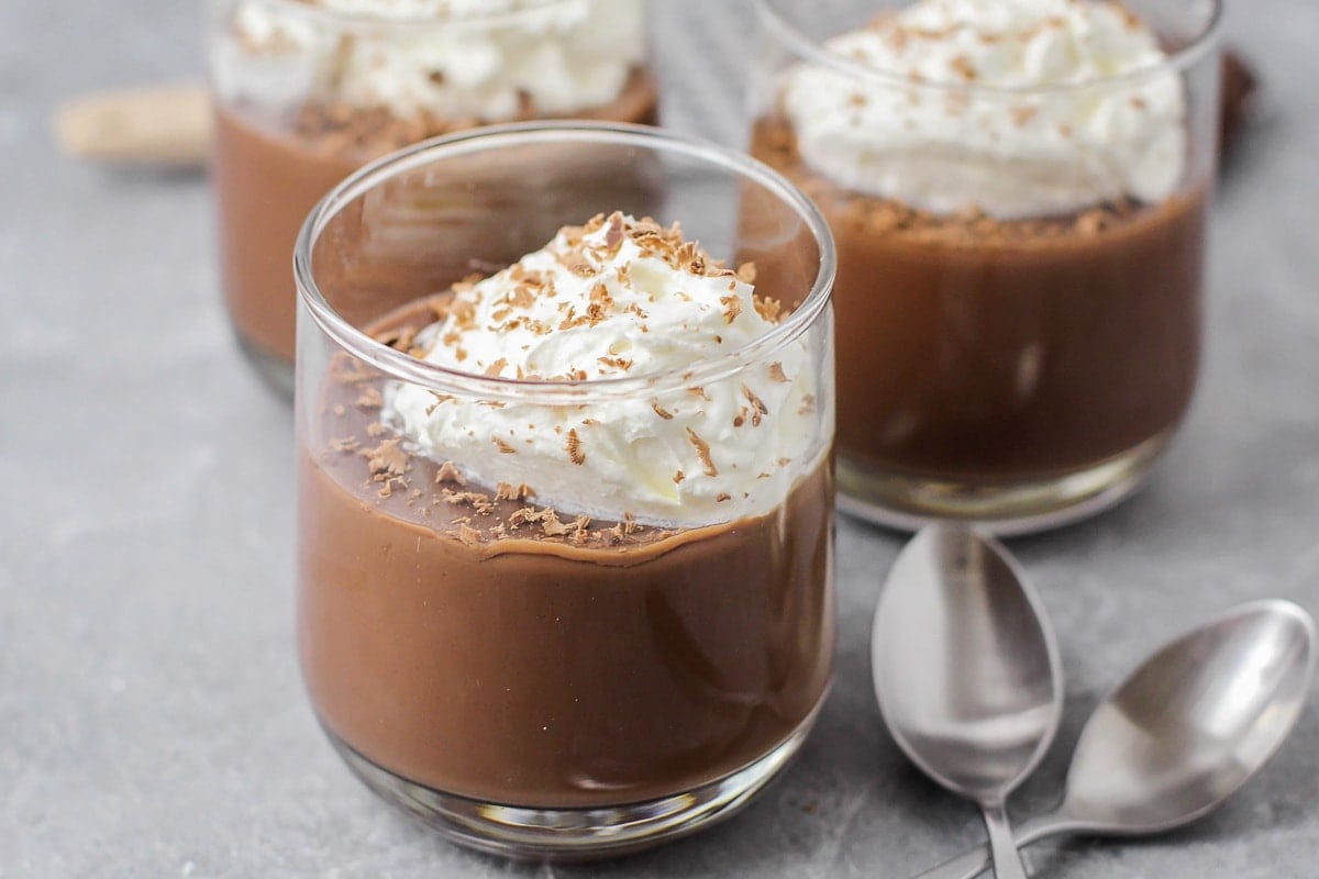 Two small glass cups filled with glossy chocolate coco pudding on a light kitchen counter, topped with a few chocolate shavings.