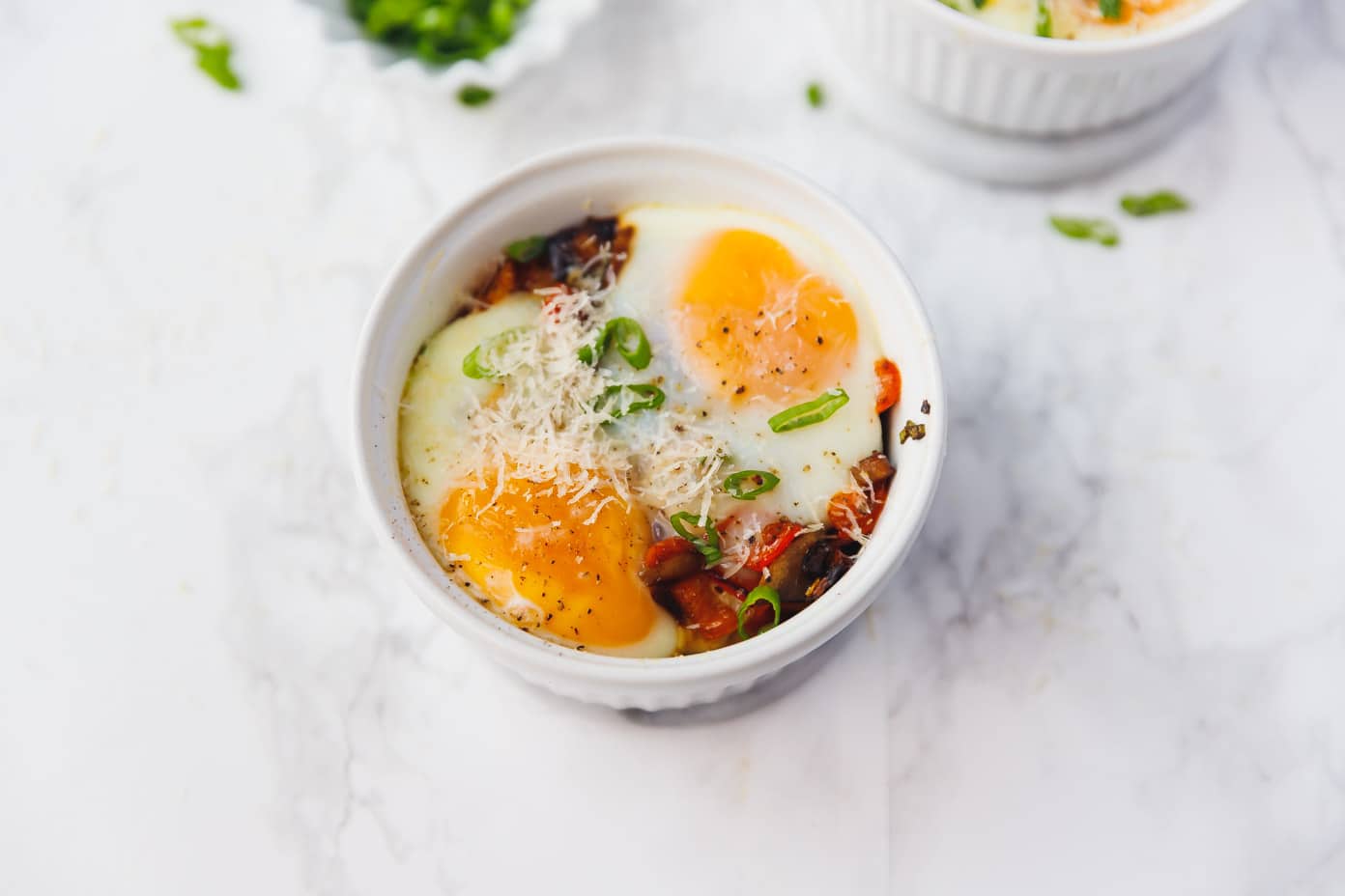 Two small ramekins of cheesy baked eggs with set whites and jammy yolks, topped with chopped chives, sitting on a light stone countertop beside toast