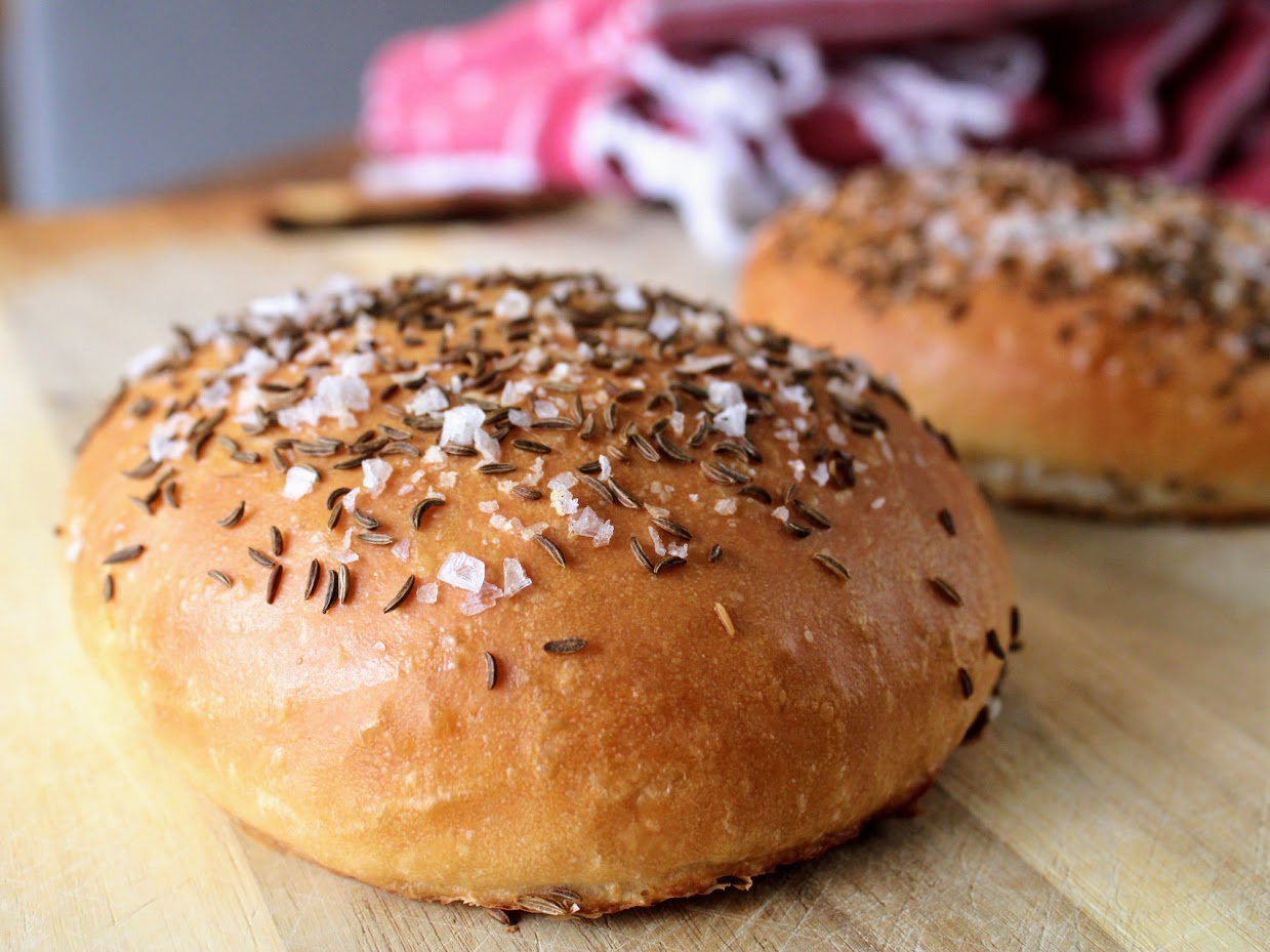 Two soft kaiser rolls on a sheet pan topped with caraway seeds and coarse salt, ready to toast, on a tidy kitchen counter