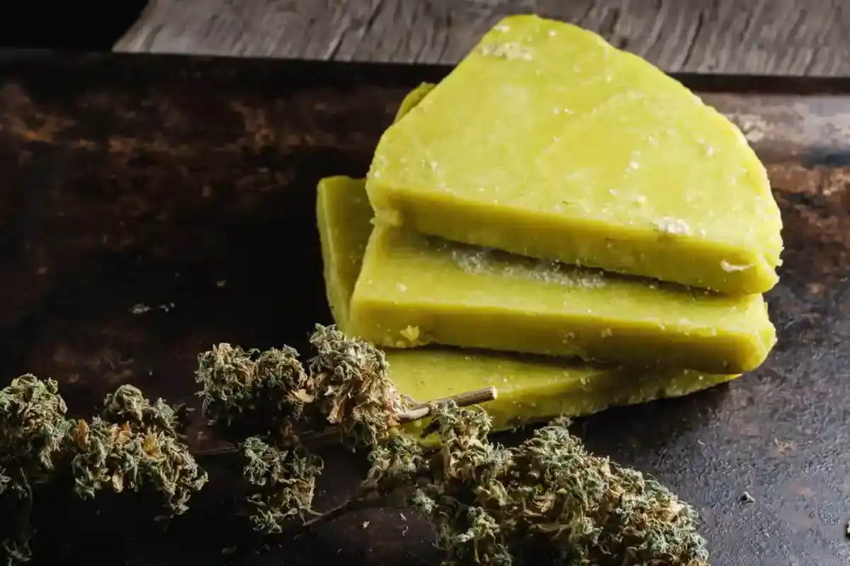 Two sticks of butter, a small bowl of cannabis flower, and a measuring cup of water set on a tidy kitchen counter with a saucepan nearby