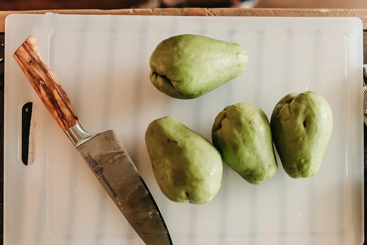 Two whole chayote squash sitting on a wooden cutting board on a clean kitchen counter with a chef's knife nearby