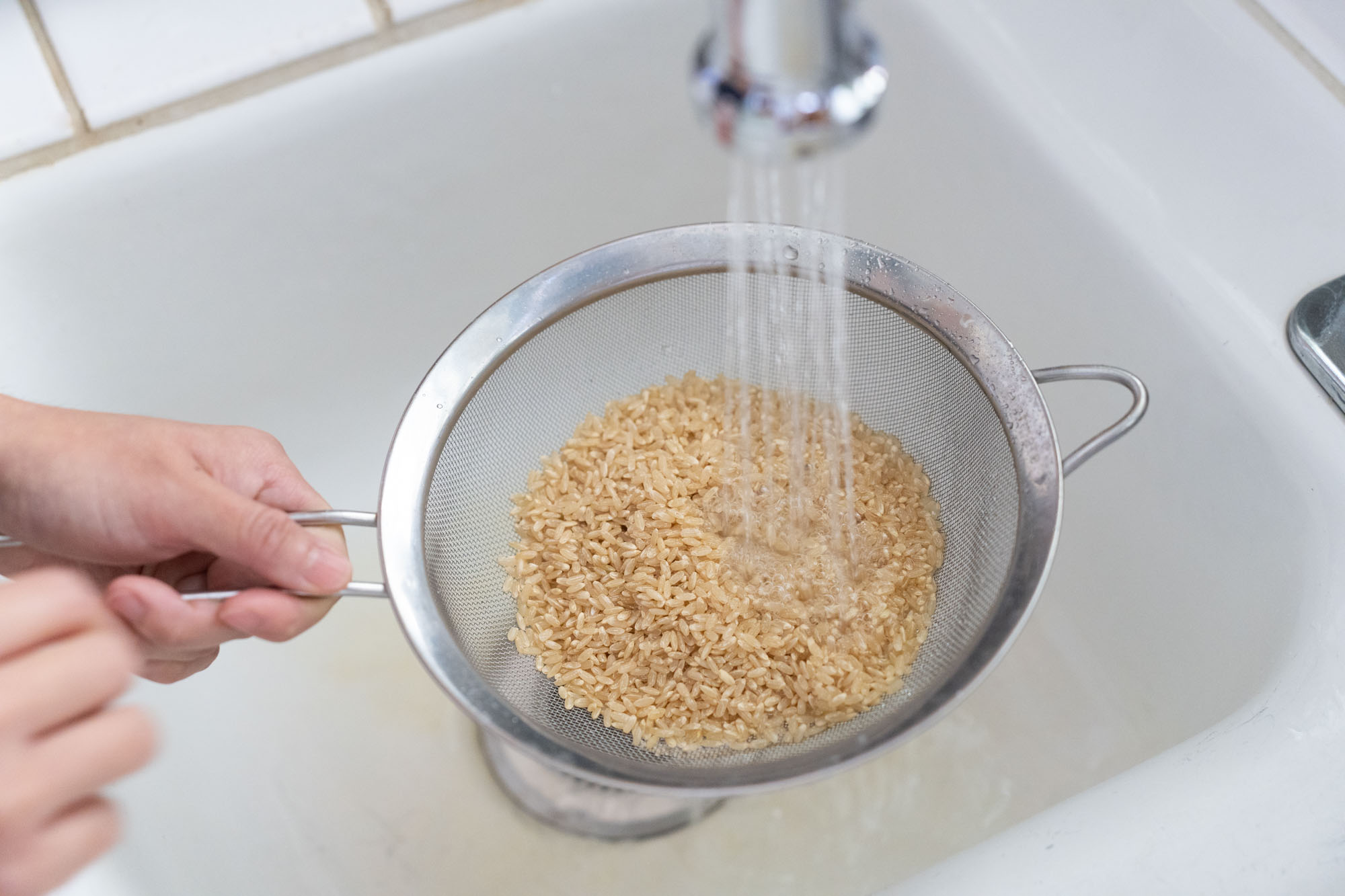 Uncooked brown rice being rinsed in a fine-mesh strainer over a sink, water running through the grains in a bright home kitchen