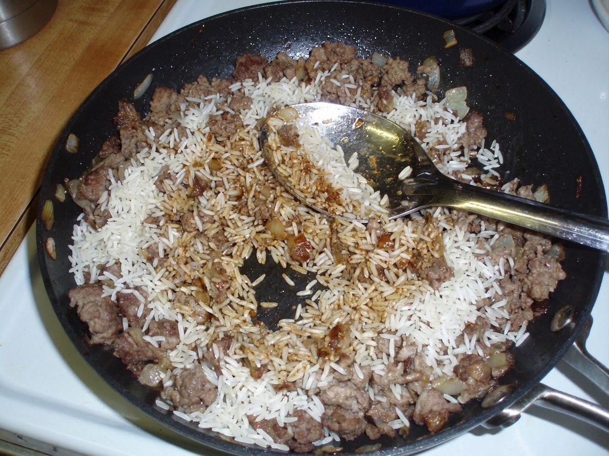 Uncooked long-grain rice being stirred into seasoned ground beef in a skillet, showing grains coated with spices