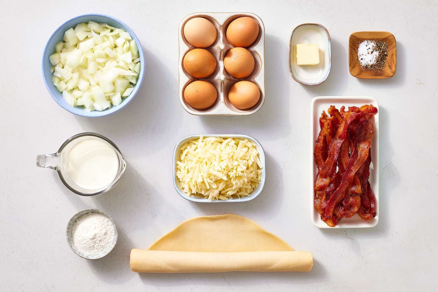 Uncooked pie crust, eggs, milk, cheddar, bacon, scallions, salt, pepper, and nutmeg arranged on a bright kitchen counter