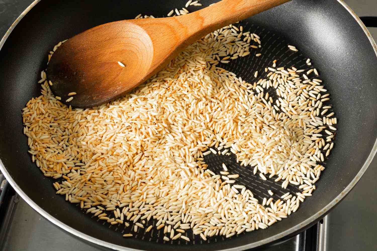 Uncooked white rice toasting in a dry skillet until lightly golden, stirred with a wooden spoon on a home stove with warm light