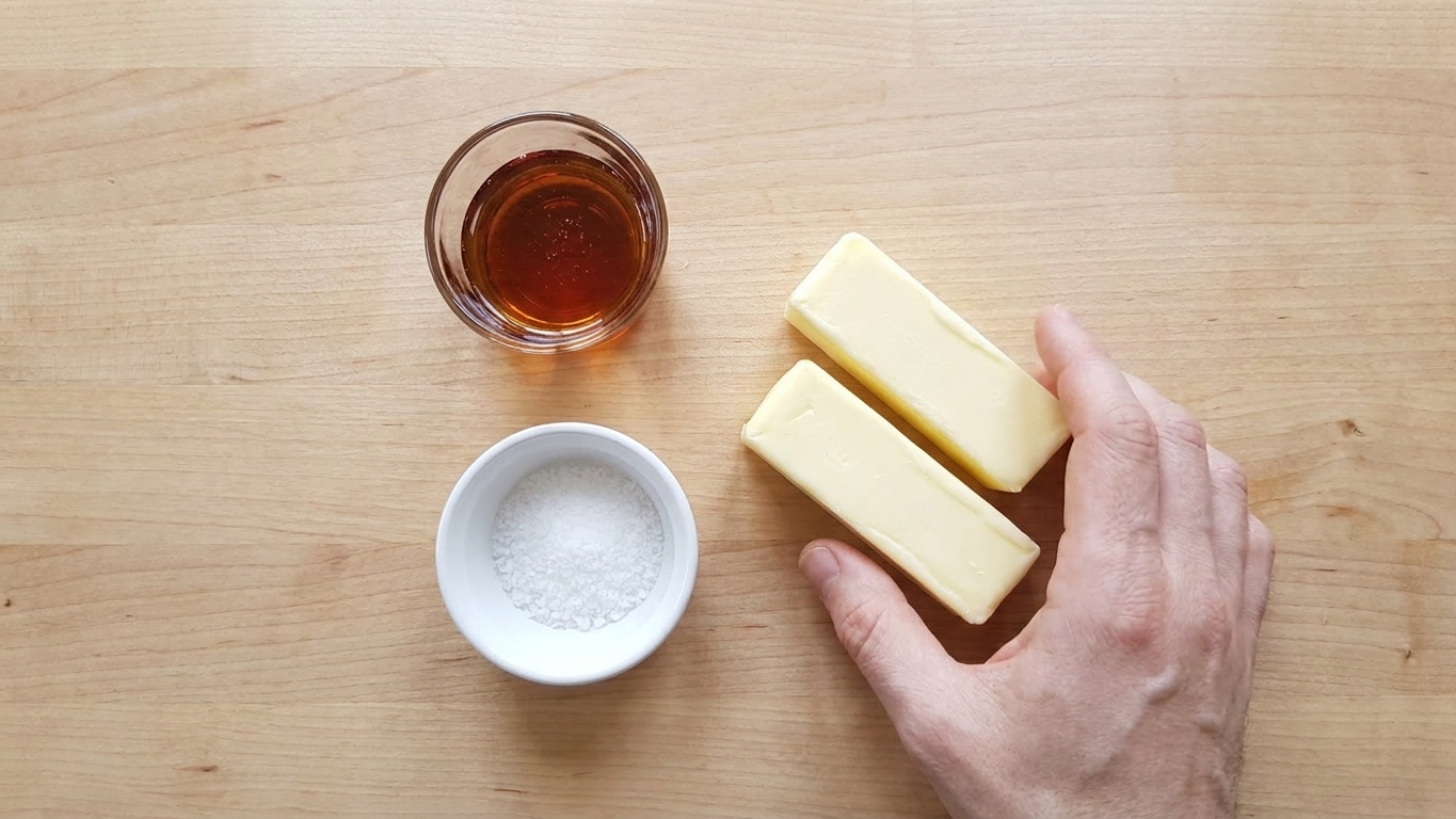 Unsalted butter, a small glass of maple syrup, and a pinch bowl of salt arranged on a light kitchen counter