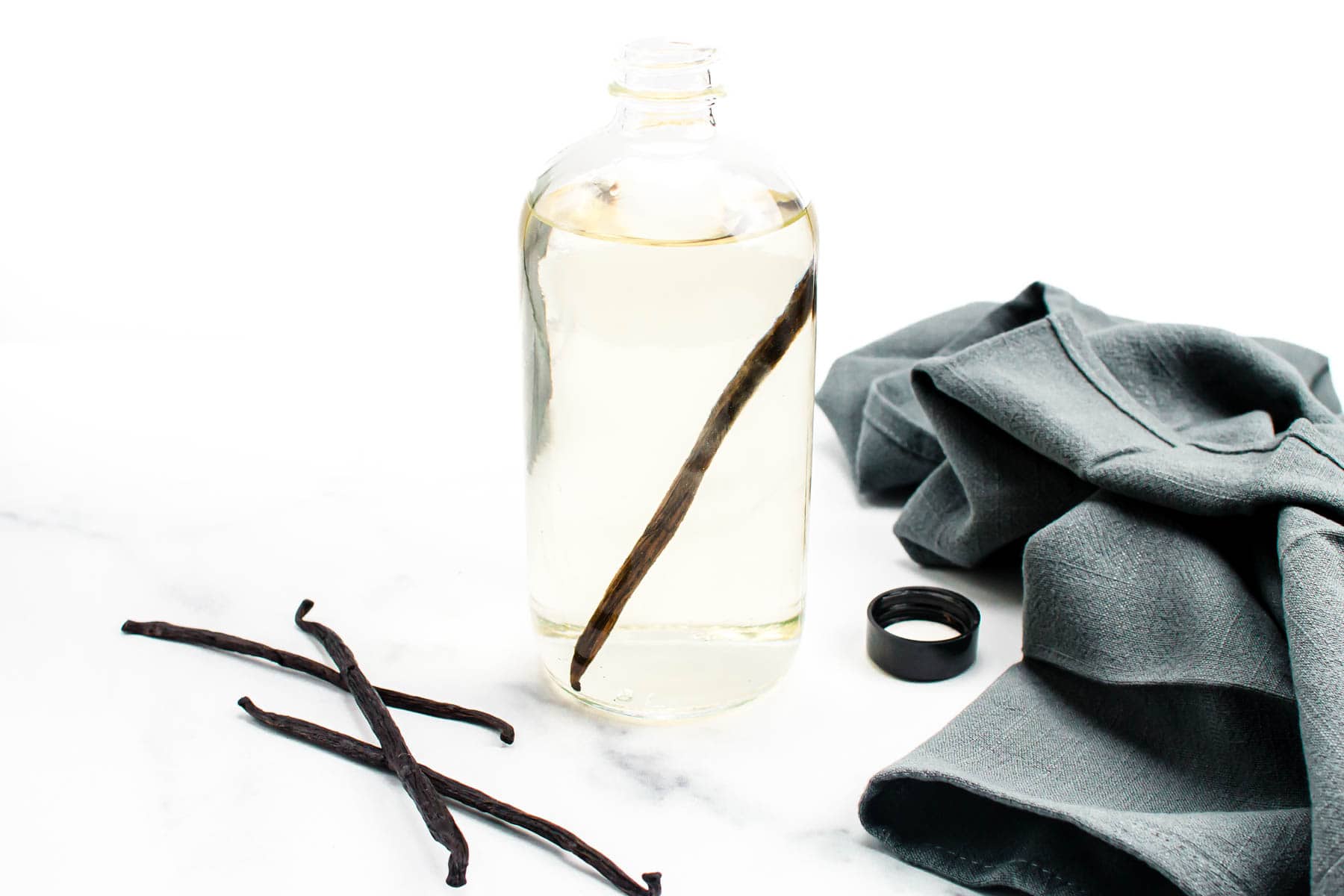 Vanilla beans, a bottle of vodka, a small funnel, and an empty glass bottle arranged on a clean kitchen counter in soft natural light