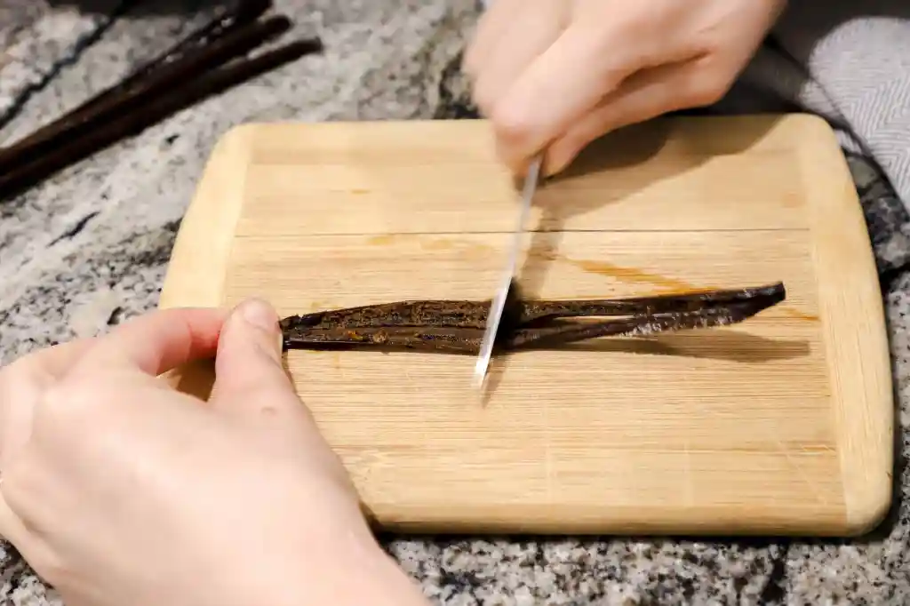 Vanilla beans being split lengthwise with a small knife on a wooden cutting board on a kitchen counter