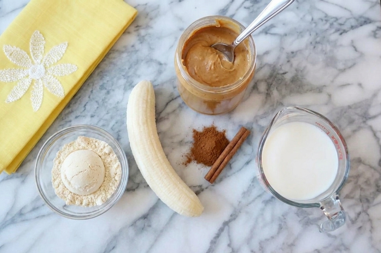 Vanilla protein powder, a peeled banana, peanut butter, cinnamon, and a measuring cup of milk arranged on a clean kitchen counter