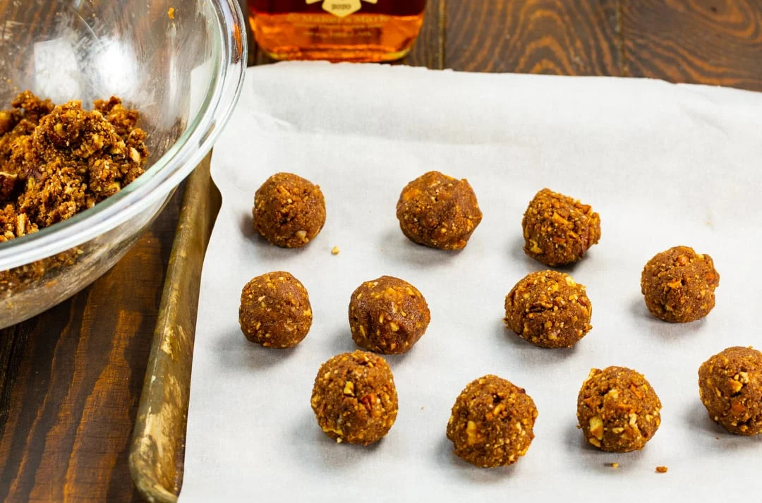 Vanilla wafer crumbs, cocoa powder, chopped toasted pecans, powdered sugar, and a small glass of bourbon arranged on a kitchen counter