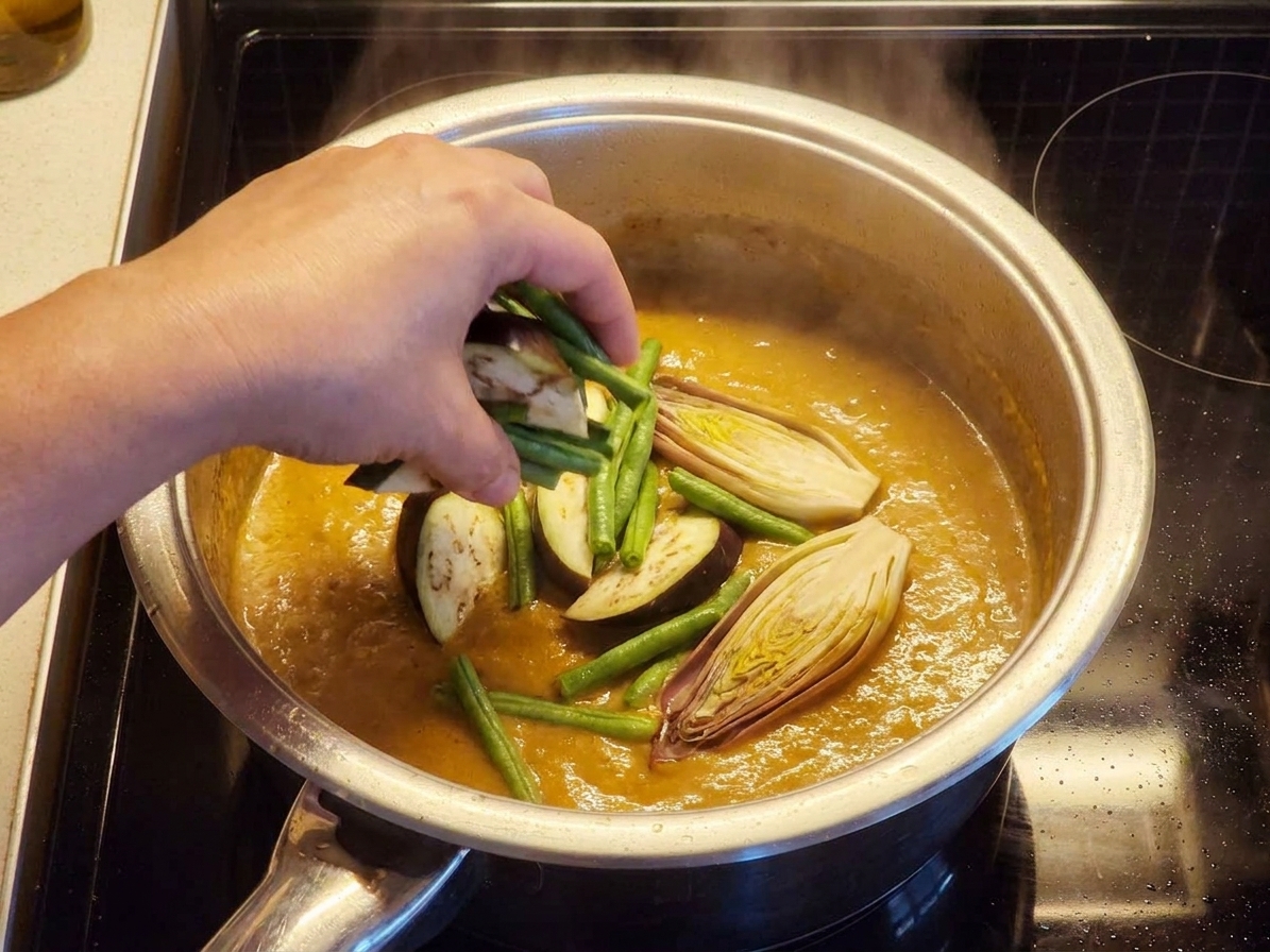 Vegetables being added into a pot of creamy peanut kare-kare sauce on the stove