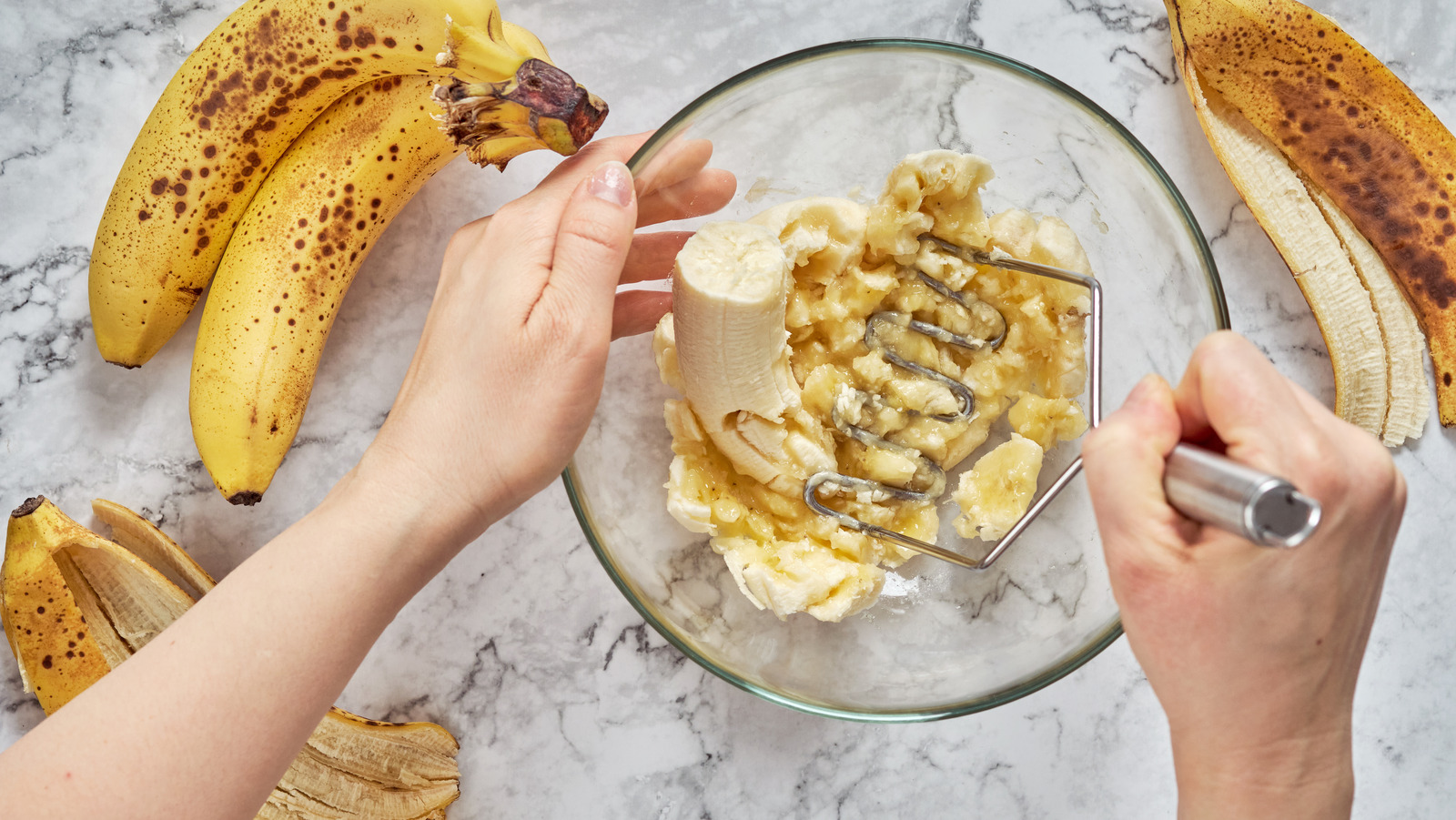 Very ripe bananas being mashed with a fork in a large mixing bowl on a kitchen counter