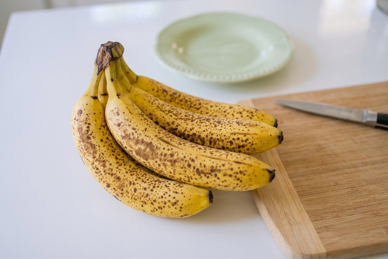 Very ripe bananas with lots of brown spots resting on a kitchen counter next to a cutting board, ready to be prepped