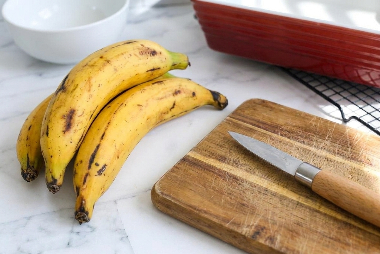 Very ripe plantains with black-speckled skins resting on a light kitchen counter next to a cutting board and a small paring knife