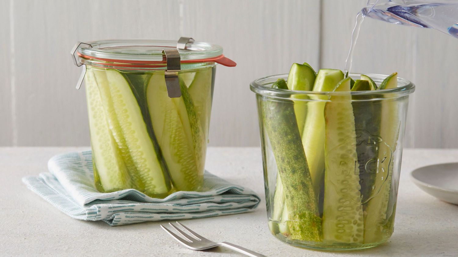 Vinegar brine being poured into a glass jar packed with sliced cucumbers, dill, and garlic on a bright kitchen counter