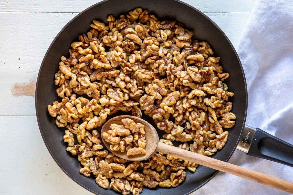 Walnuts toasting in a dry skillet on a stovetop, lightly browned and fragrant, with a wooden spoon nearby