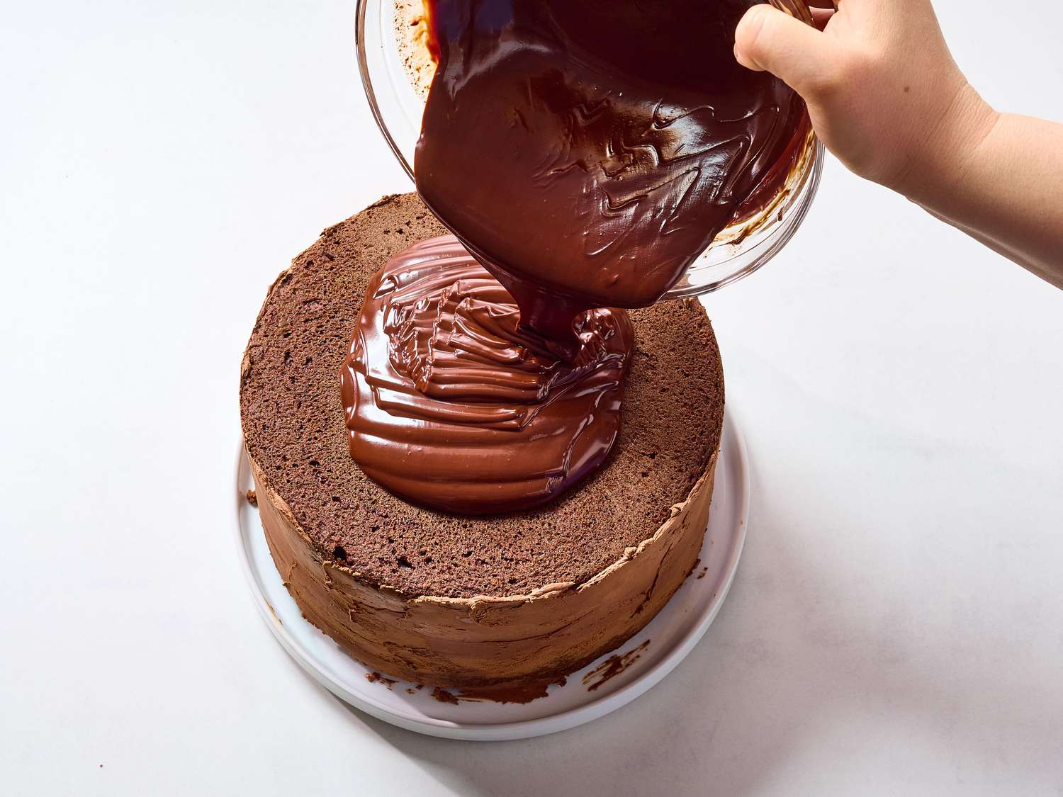 Warm chocolate frosting being poured from a saucepan over a freshly baked chocolate sheet cake in a baking pan