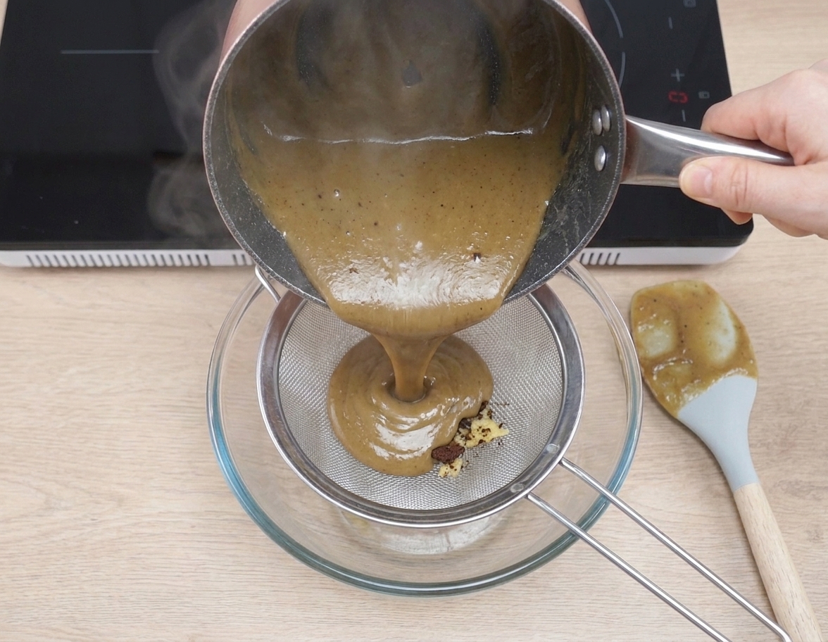 Warm coffee custard being poured through a fine-mesh strainer into a bowl on a kitchen counter