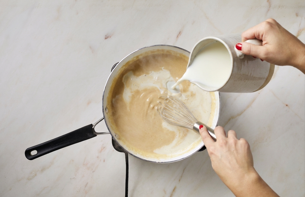 Warm milk being poured into a saucepan while whisking to create a smooth béchamel base