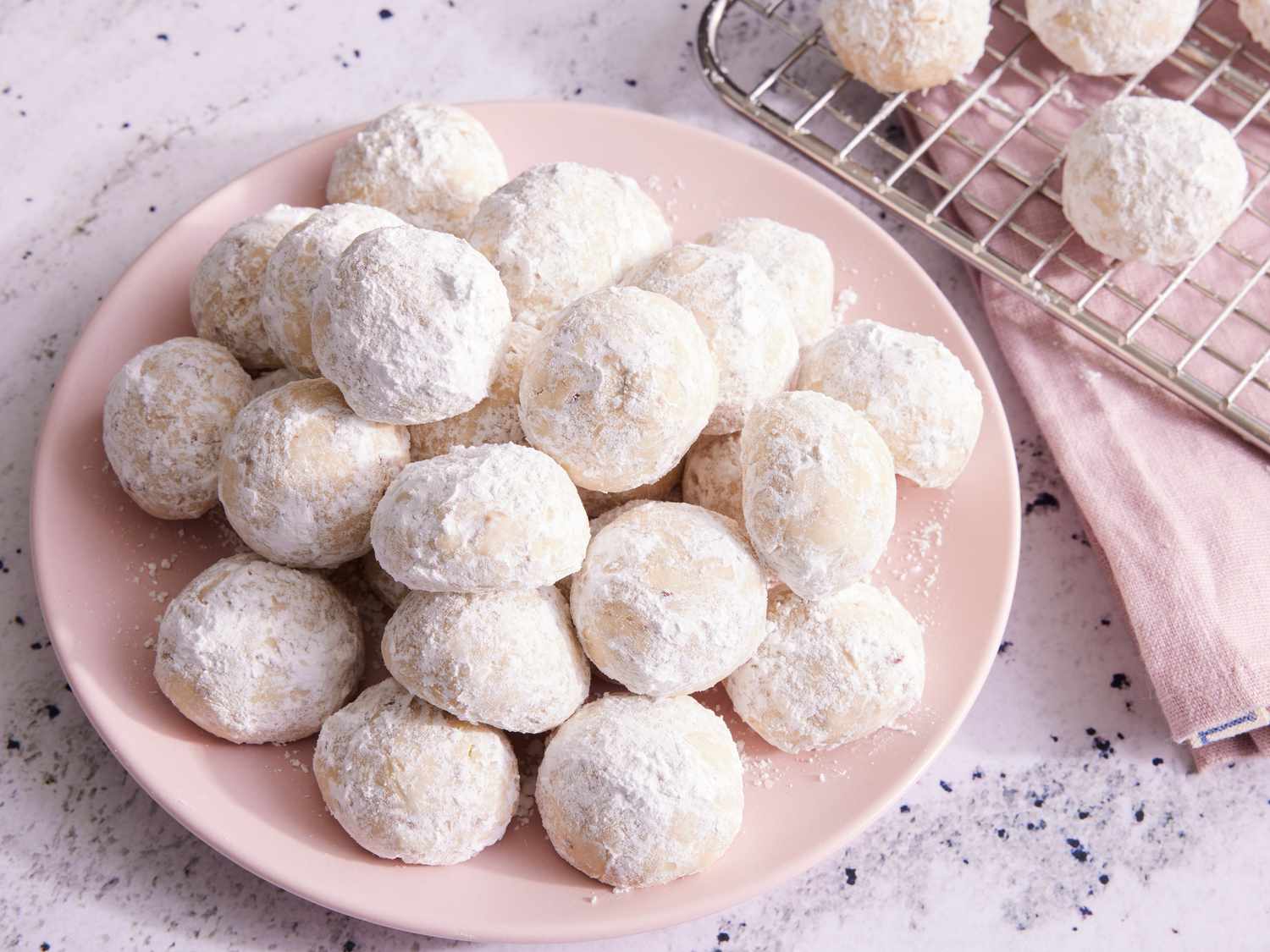 Warm snowball cookies being gently rolled in a shallow bowl of powdered sugar on a kitchen counter with a parchment-lined baking sheet nearby