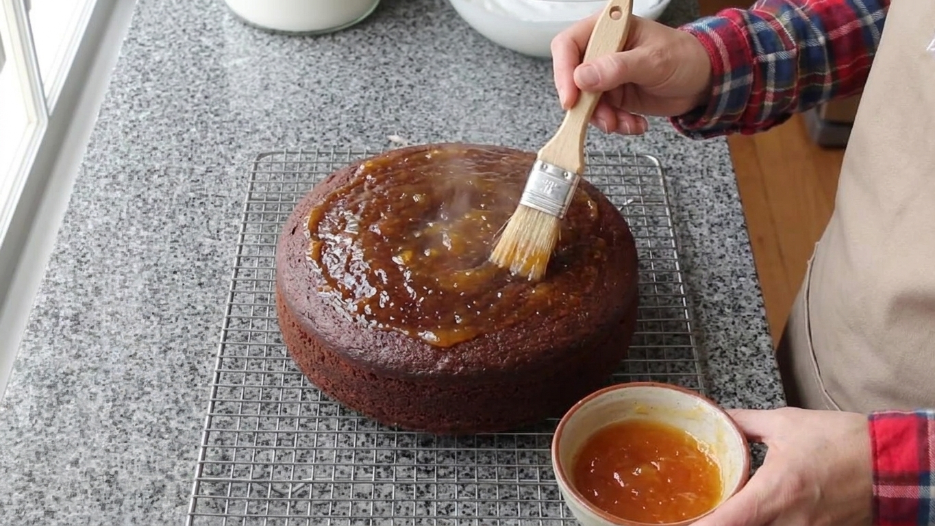 Warm strained apricot jam being brushed over the top of a chocolate cake on a wire rack