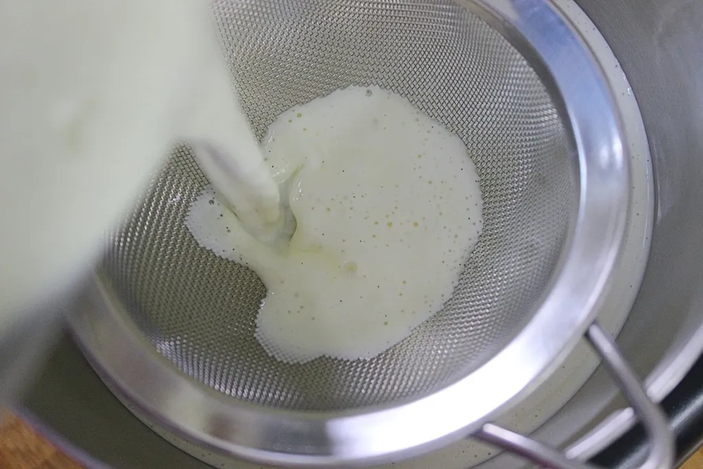 Warm vanilla custard being poured through a fine-mesh sieve into a bowl on a kitchen counter, with a whisk and saucepan nearby