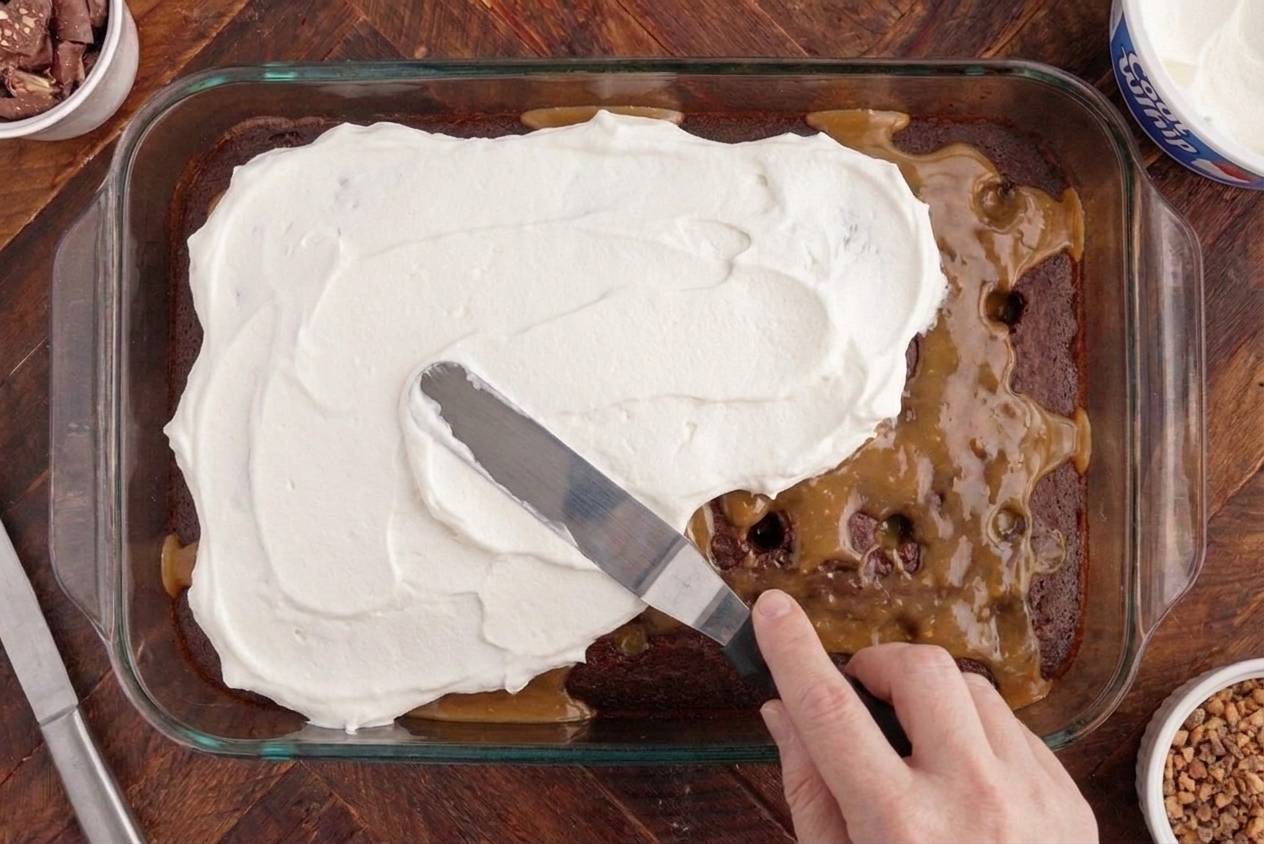 Whipped topping being spread evenly over a chilled chocolate poke cake in a 9x13 pan using an offset spatula