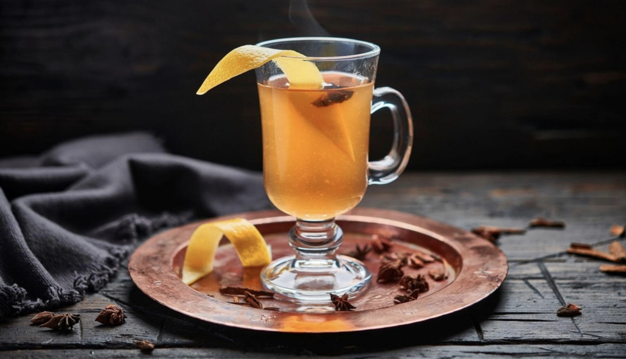 Whiskey, a lemon, a jar of honey, cinnamon sticks, and a kettle arranged on a tidy kitchen counter under warm light