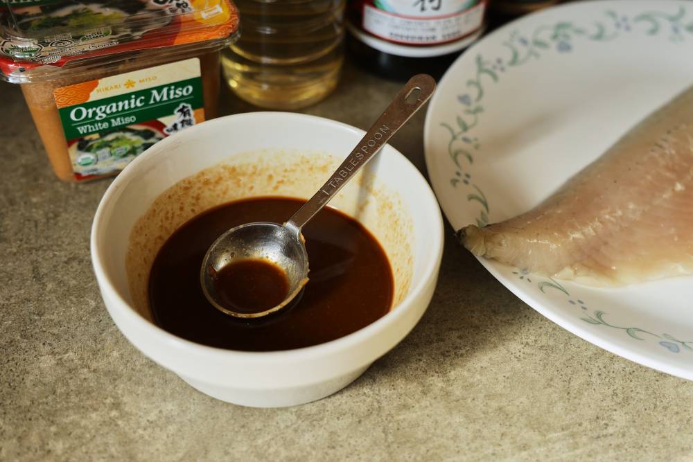 White miso paste, mirin, soy sauce, brown sugar, and fresh ginger measured on a kitchen counter next to a small mixing bowl
