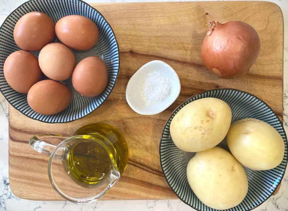 Whole Yukon Gold potatoes, a yellow onion, eggs, and a bottle of olive oil arranged on a clean kitchen counter with soft natural light