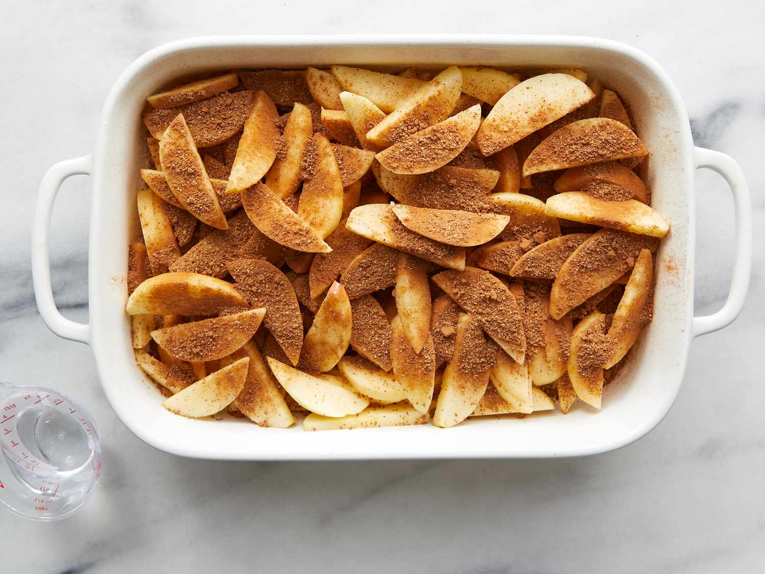 Whole apples, a small bowl of cinnamon, a lemon, brown sugar, and a measuring cup on a light kitchen counter