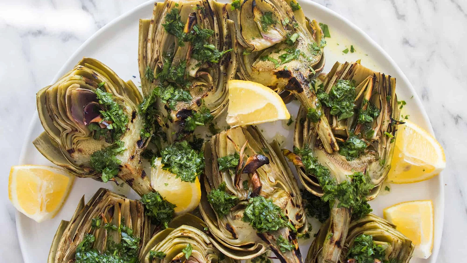 Whole fresh globe artichokes on a cutting board with lemons, garlic, parsley, olive oil, and a bowl of breadcrumbs on a kitchen counter