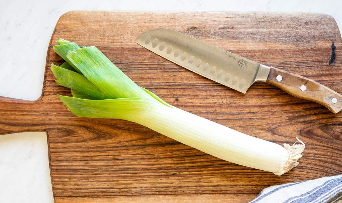 Whole fresh leeks with roots trimmed sitting on a wooden cutting board beside a chef's knife and a small bowl, in a bright home kitchen