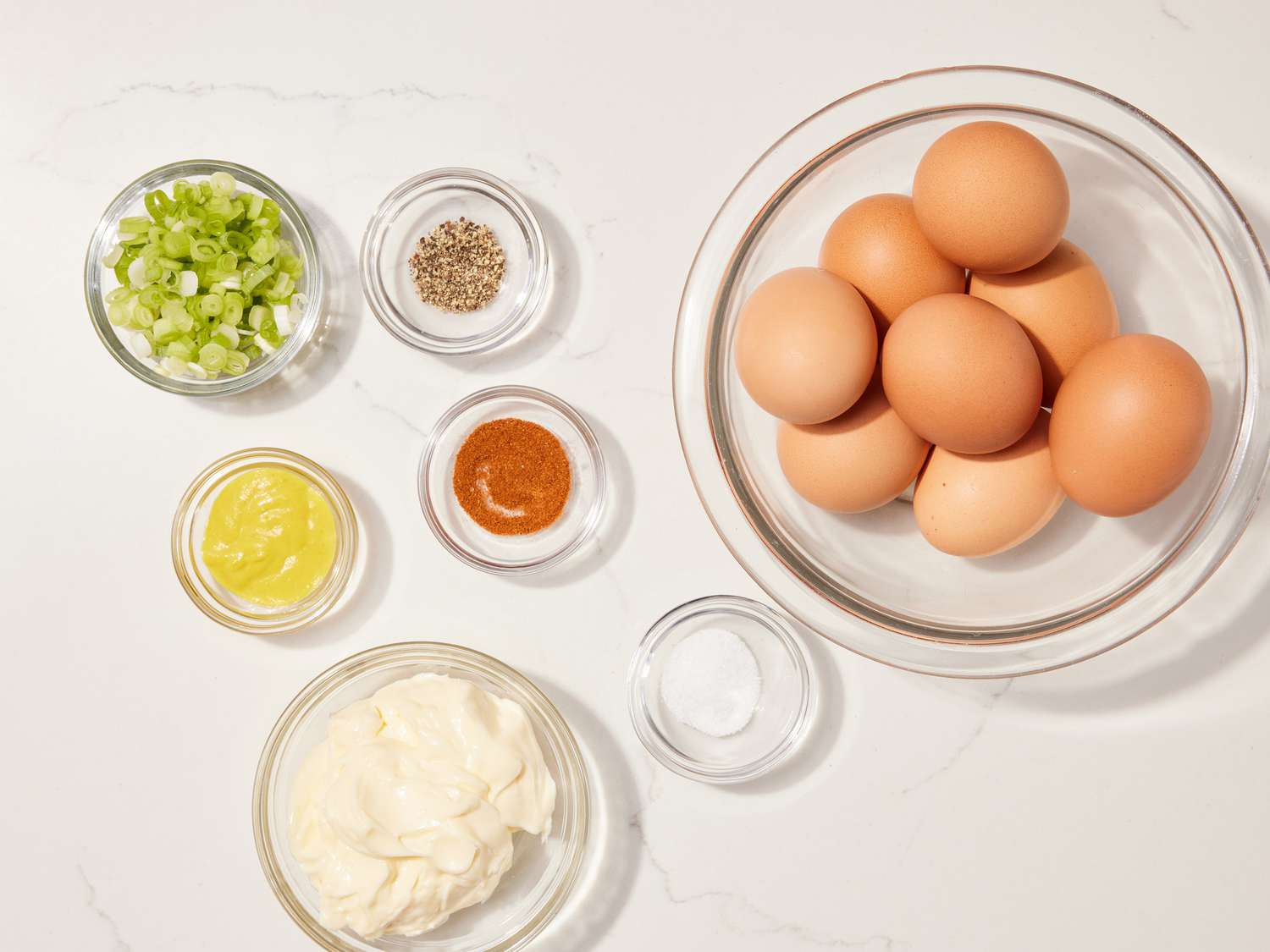 Whole ingredients for egg salad including hard-boiled eggs, mayonnaise, Dijon mustard, lemon, celery, red onion, fresh dill, salt, and pepper arranged on a kitchen counter