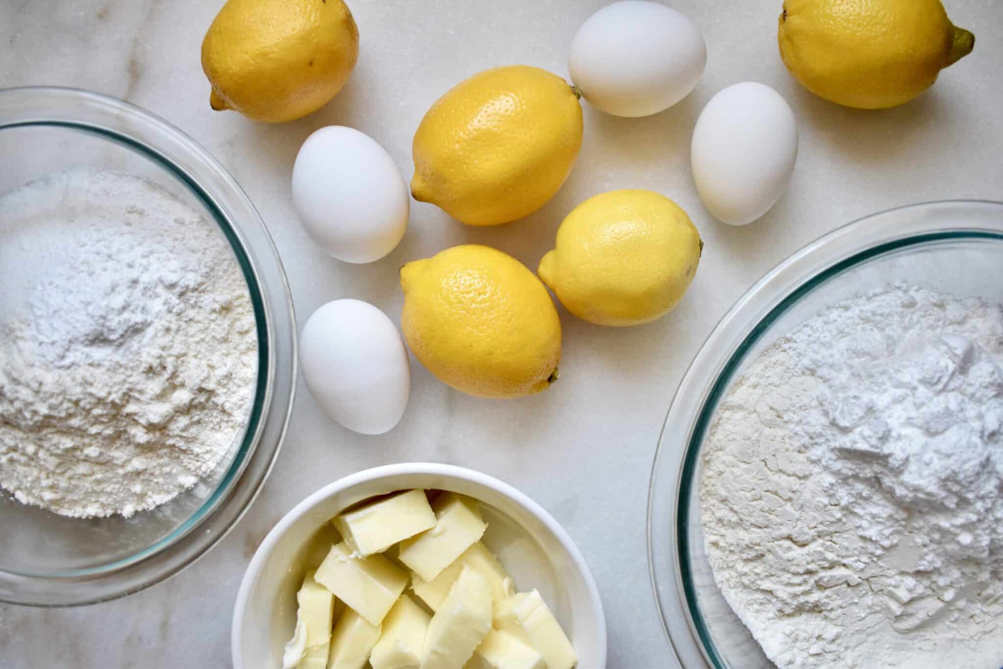 Whole lemons, butter, eggs, flour, and sugar arranged on a clean kitchen counter in warm natural light