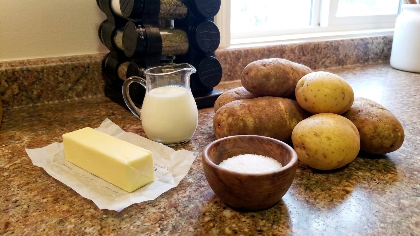 Whole russet and Yukon Gold potatoes on a bright kitchen counter next to kosher salt, butter, and a small pitcher of cream
