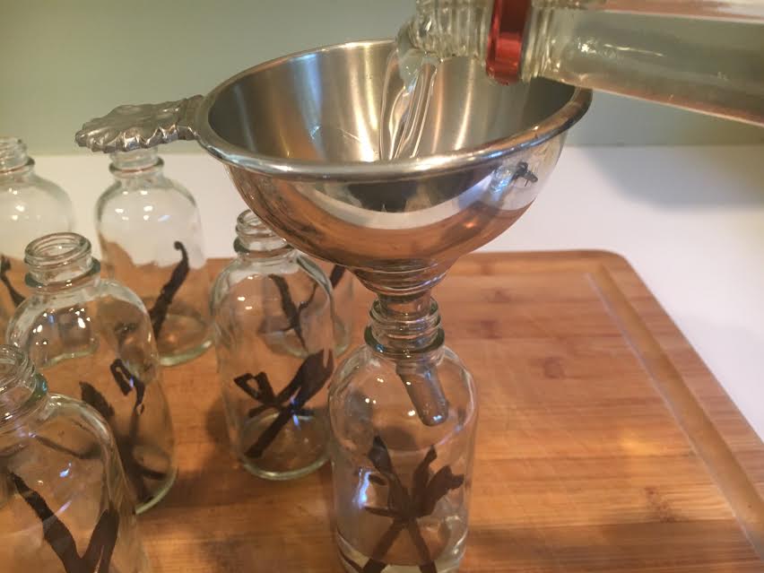 Whole vanilla beans, a bottle of vodka, an amber jar, and a small stainless steel funnel arranged on a kitchen counter