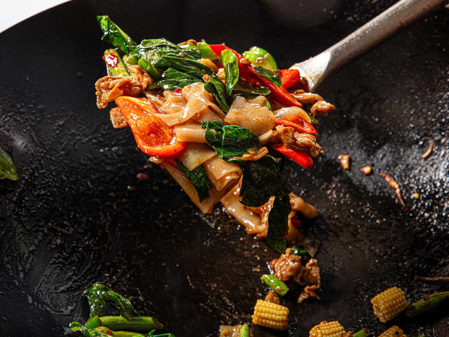 Wide rice noodles being tossed in a hot wok with tongs over a home stovetop